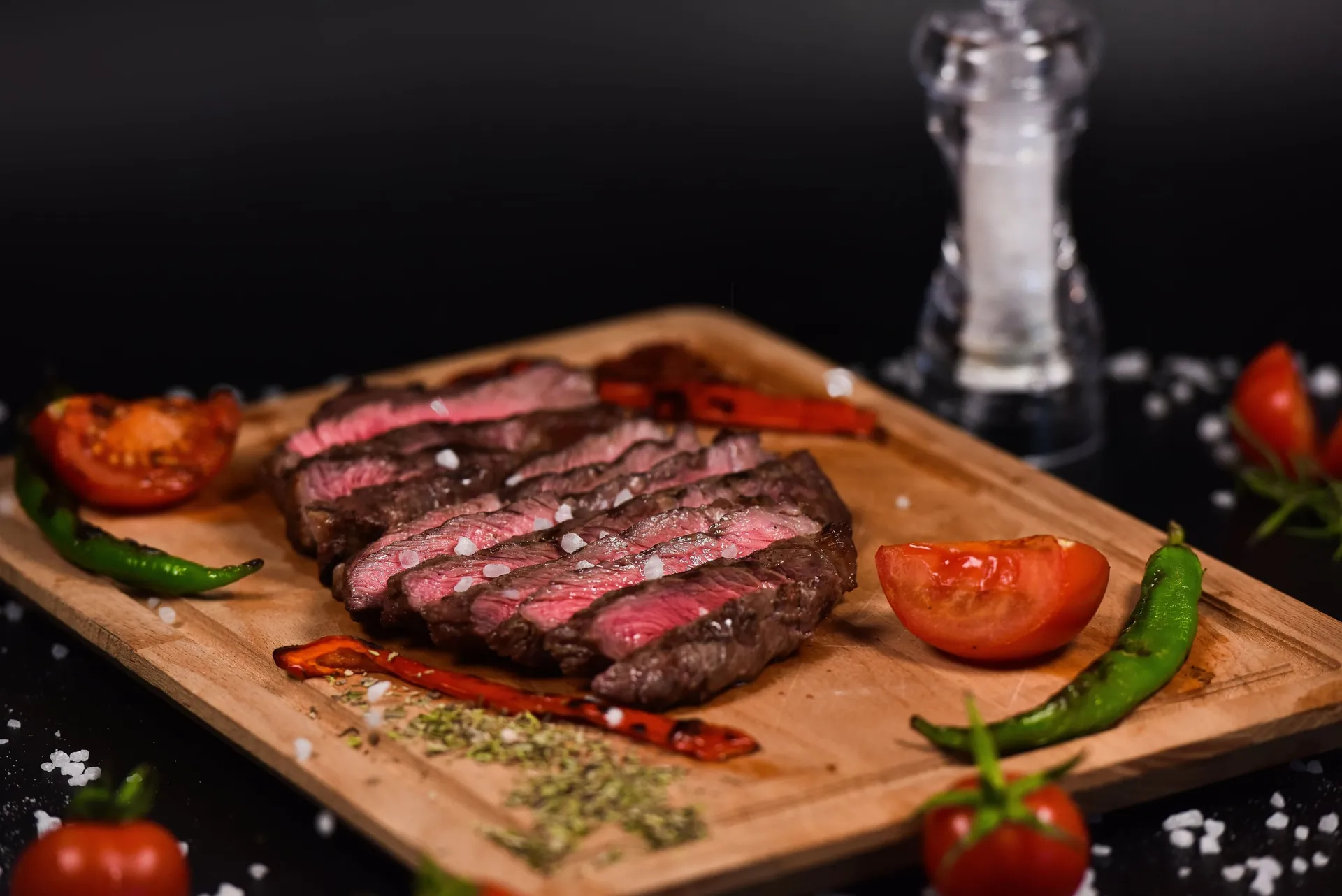 Gesneden rosé gegrilde biefstuk met grof zout, tomaat en groene pepers op een houten plank